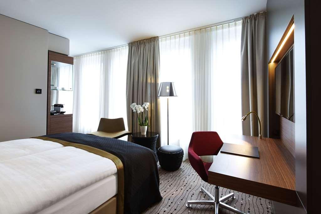 Steigenberger Hotel Am Kanzleramt Berlin Room photo The photo shows a modern hotel room featuring a bed with a white comforter and a dark accent blanket. On one side of the room, there are large windows with sheer curtains allowing natural light to enter. A small desk with a lamp is set up near the wi