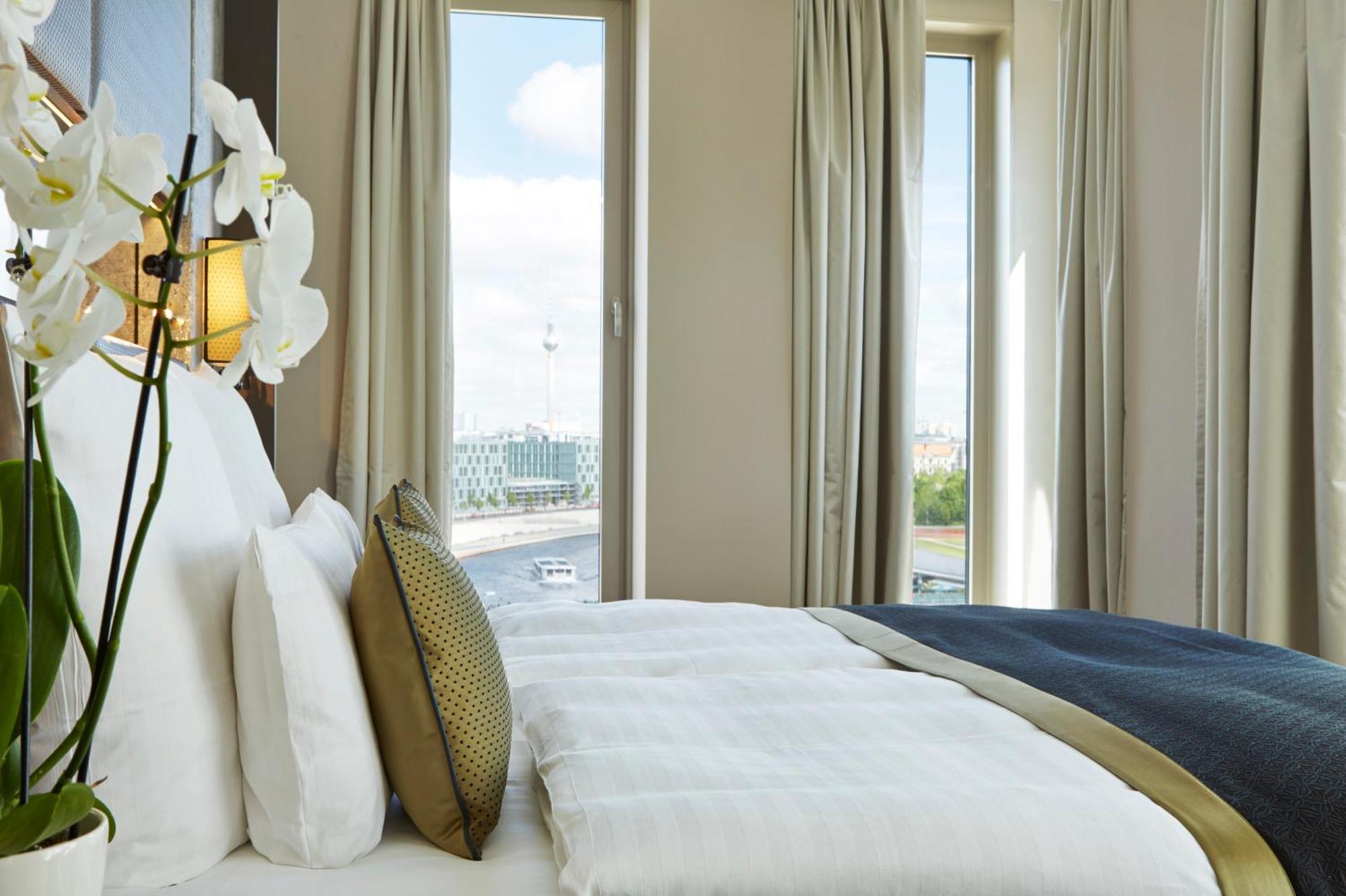 Steigenberger Hotel Am Kanzleramt Berlin Room photo The photo shows a well-decorated hotel room with a large bed featuring plush white bedding and decorative pillows. There are elegant curtains framing large windows that allow natural light to enter the space. In the foreground, a vase with white orch