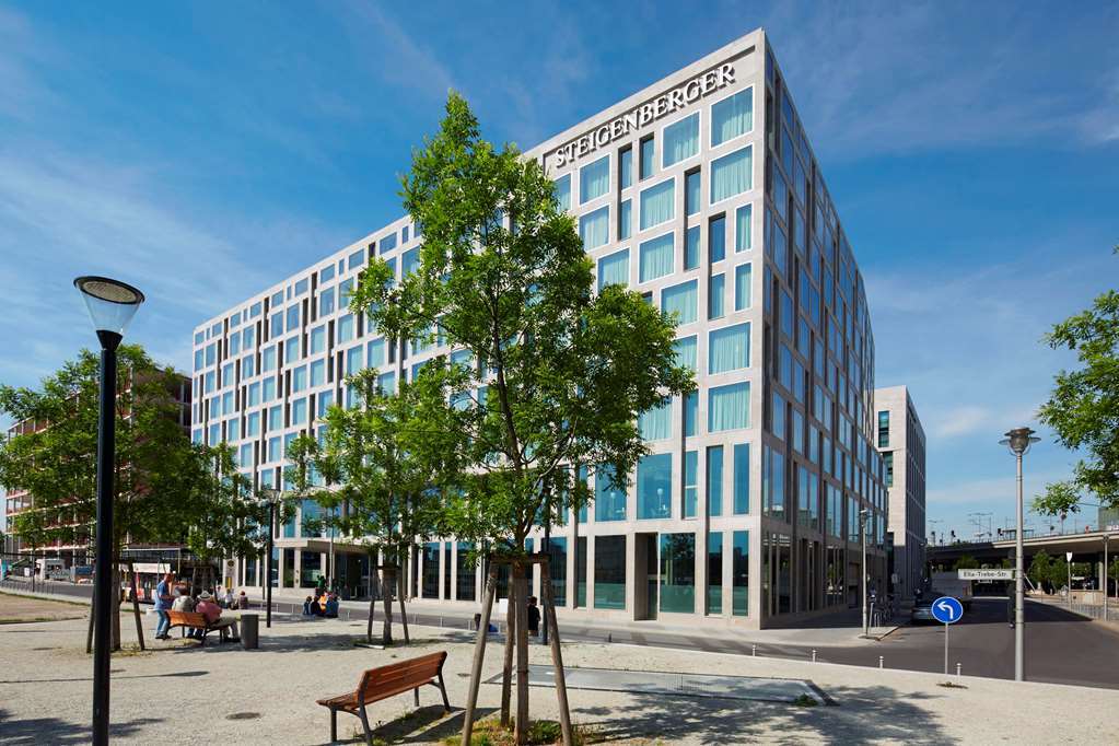 Steigenberger Hotel Am Kanzleramt Berlin Exterior photo The photo shows a modern, multi-story building with a large glass facade, featuring a grid pattern of windows. The building has the name "Steigenberger" prominently displayed at the top. Surrounding the structure are green trees and a landscaped area