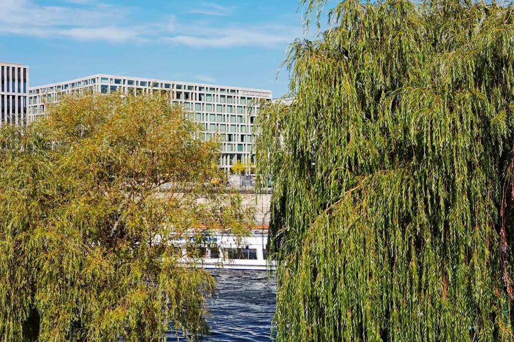 Steigenberger Hotel Am Kanzleramt Berlin Exterior photo The photo shows a view with lush, green willows in the foreground, characterized by their long, drooping branches. In the background, there is a modern building with large windows, suggesting a contemporary architectural style. The scene also include