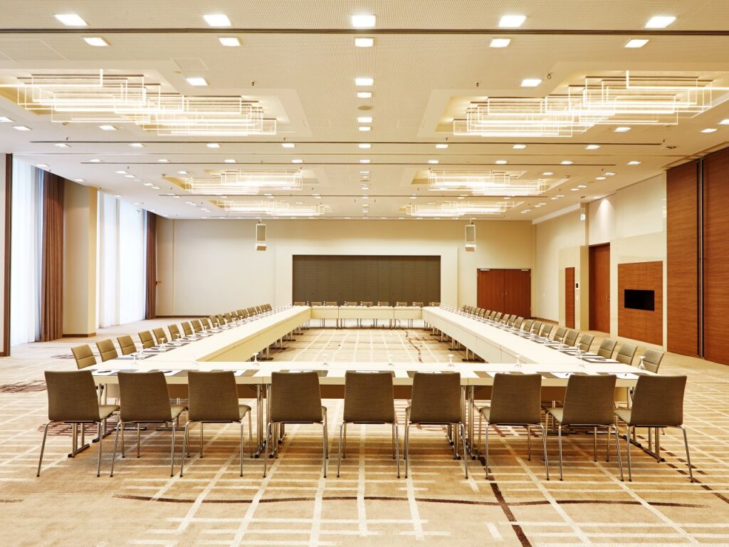 Steigenberger Hotel Am Kanzleramt Berlin Exterior photo The photo shows a large, modern conference room designed for meetings or presentations. It features a long, rectangular table set up in the center, surrounded by multiple chairs. The room is well-lit with contemporary lighting fixtures on the ceiling